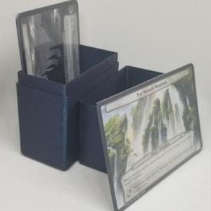 Once you have acquired you Planechase sleeves, you will need a deckbox to hold them all. Shown in the photo is a sparkly blue 3D printed deckbox for oversized MTG sleeves. The deckbox is open showing a few cards sitting inside it and once card outside of it, leaned up against the top half of the deckbox.